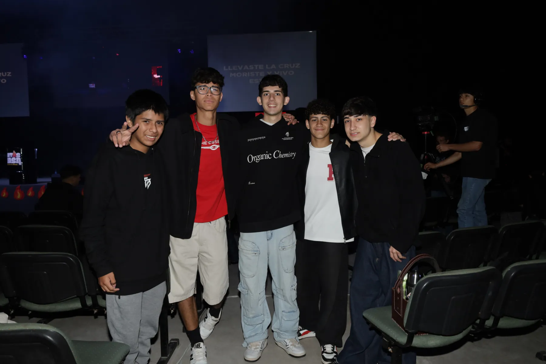 Un grupo de chicos posando y sonriendo entre las butacas, con las pantallas del escenario detrás.